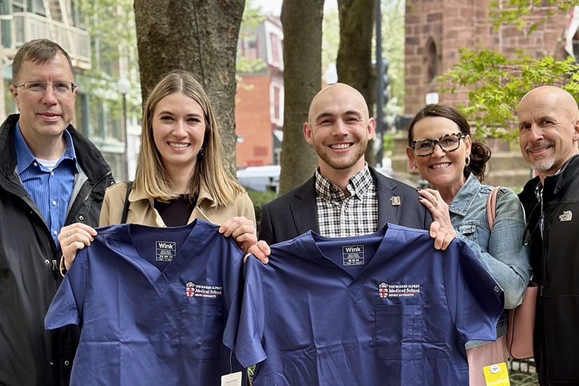 A female and male medical student from the class of 2027 hold up blue shirts with The Warren Alpert Medical School's logo, with three other people standing behind them outdoors.