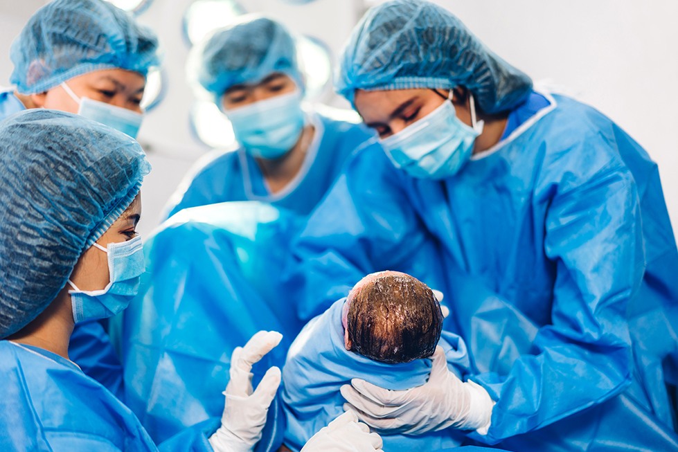 A group of doctors in blue scrubs wearing medical masks and caps examine a newborn baby in a white hospital room.
